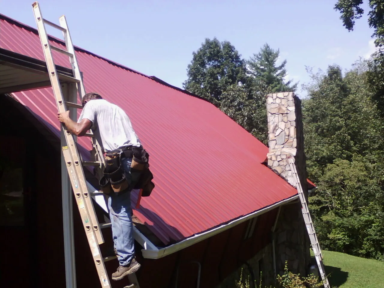 Roofer on a ladder beside a red metal roof and stone chimney — Soffit & Fascia Repair by Roofing Brothers in Lucedale